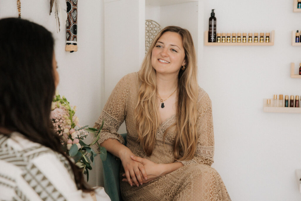 Zwei Frauen sitzend sich lächelnd gegenüber, ätherische Öle im Hintergrund
