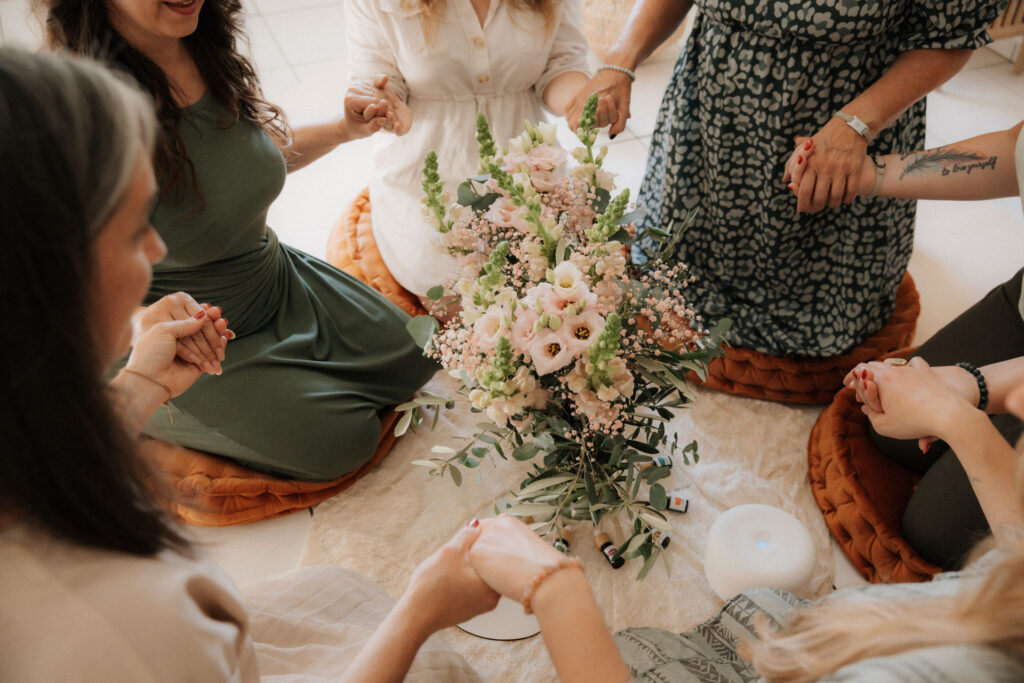 Mehrere Frauen sitzen im Kreis und halten sich die Hände in der Mitte liegen ätherische Öle und ein Blumenstrauss